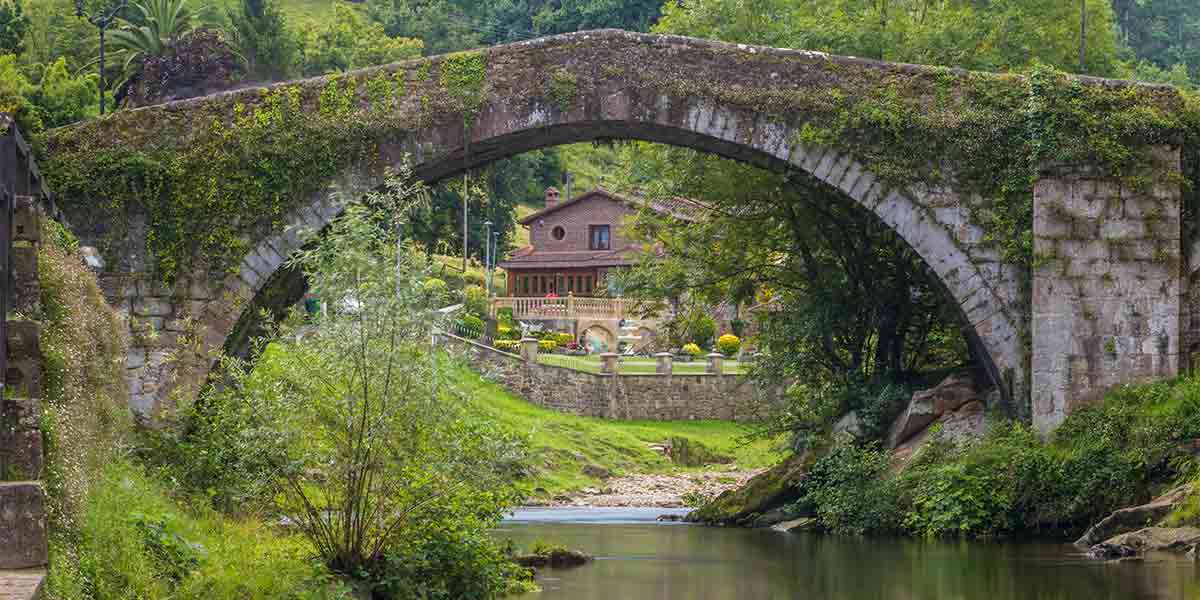 Un pintoresco escenario de Liérganes, unos de los pueblos con más encanto de Cantabria, donde se ve un antiguo puente de piedra sobre el río, rodeado de vegetación y recubierto por un manto de plantas verdes