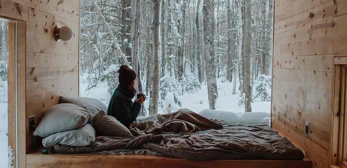 Una mujer relajada metida en la cama de una cabaña del bosque con un café en las manos