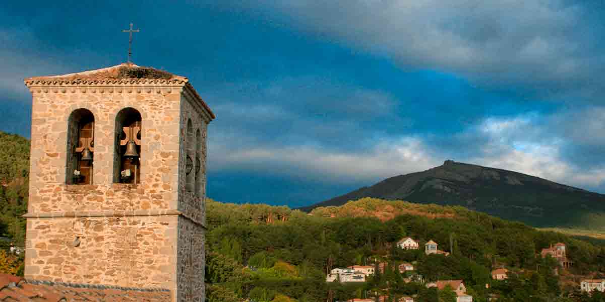 La histórica torre de la Iglesia Parroquial de la Asunción de Nuestra Señora de Miraflores de la Sierra con las verdes montañas por detrás y la bonita luz del atardecer