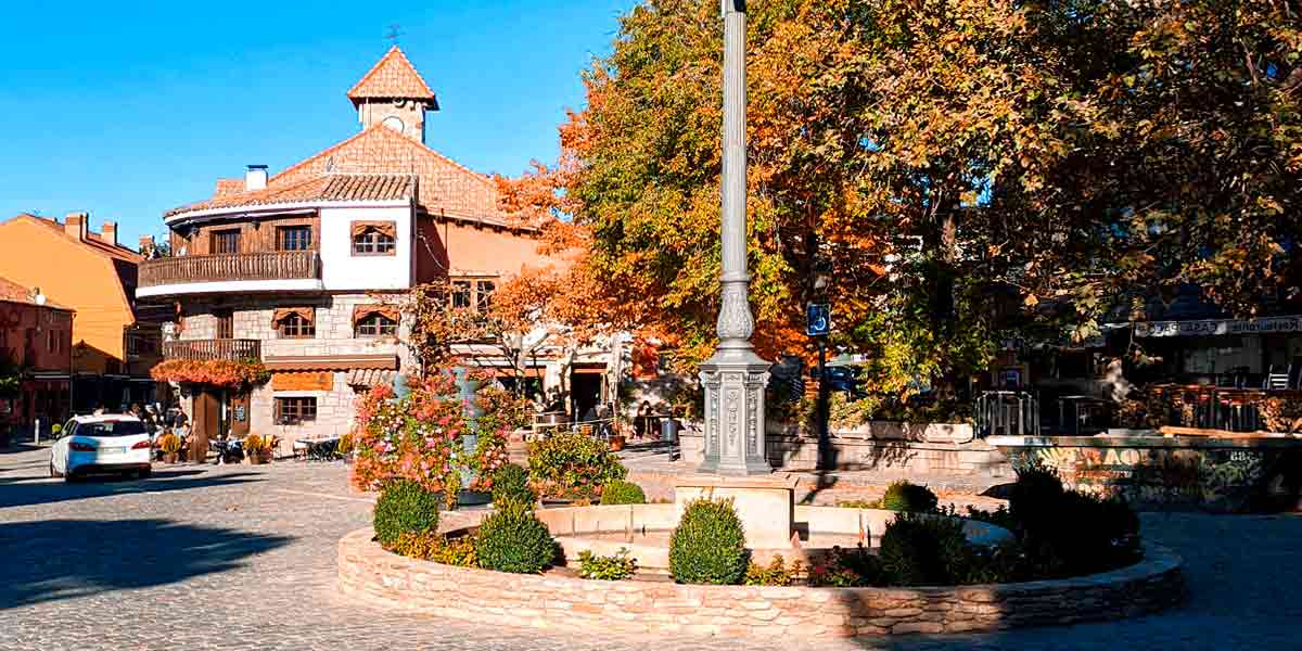 La preciosa plaza principal de Navacerrada repleta de árboles con hojas verdes y naranjas por el otoño y con un precioso y despejado cielo azul