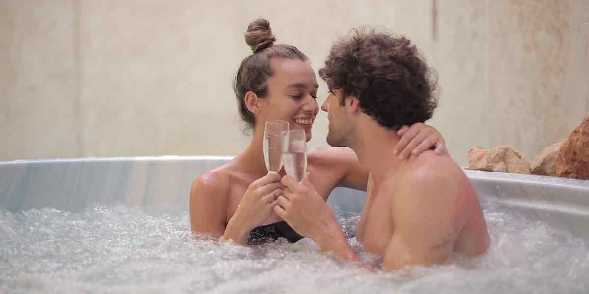 Una pareja en un hotel con jacuzzi en la habitación sumergidos en el burbujeante agua mientras celebran un momento especial brindando con copas de champagne